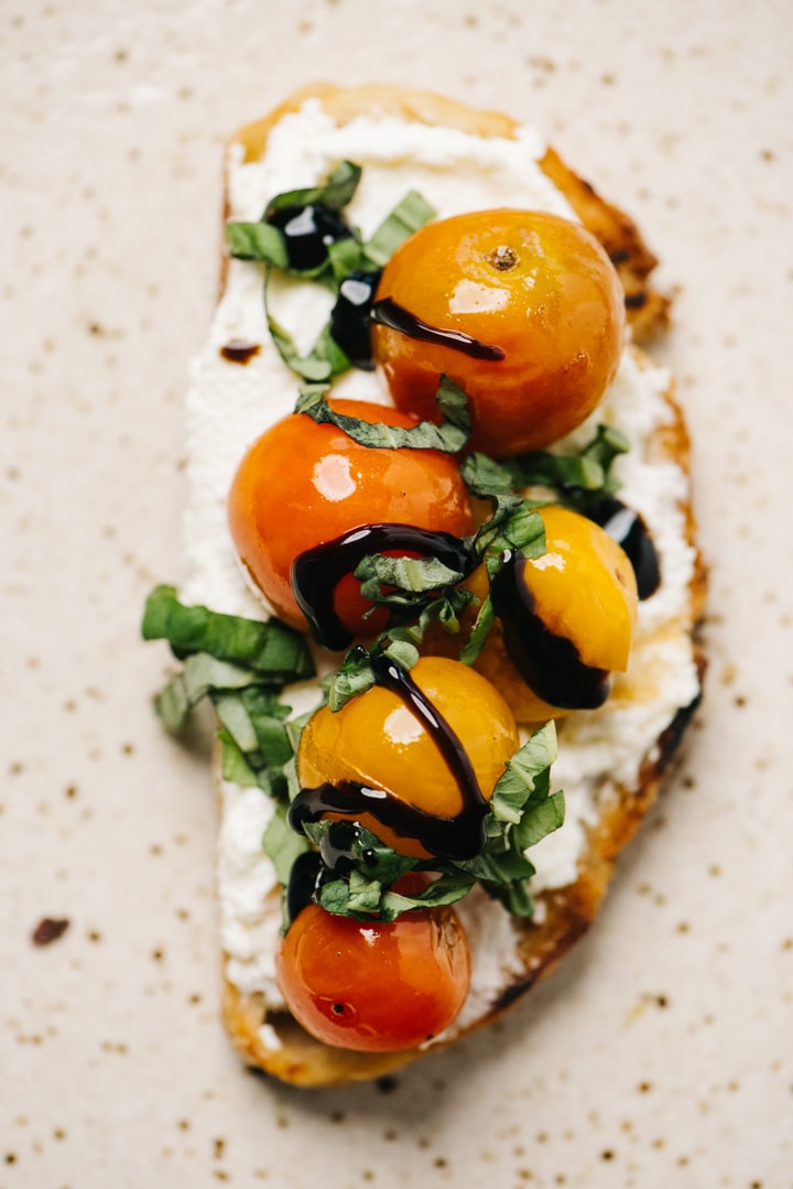 A slice of savory ricotta toast topped with burst tomatoes, fresh basil, and balsamic glaze on a speckled tan plate.