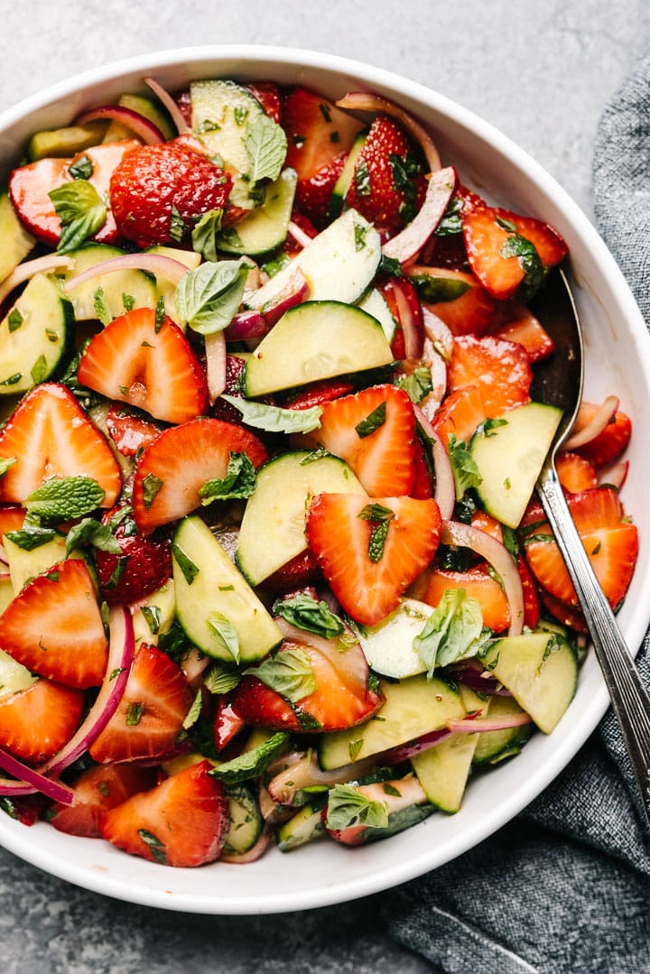 Strawberry cucumber salad with red onions in a white serving bowl with a vintage serving spoon and grey linen napkin to the side.