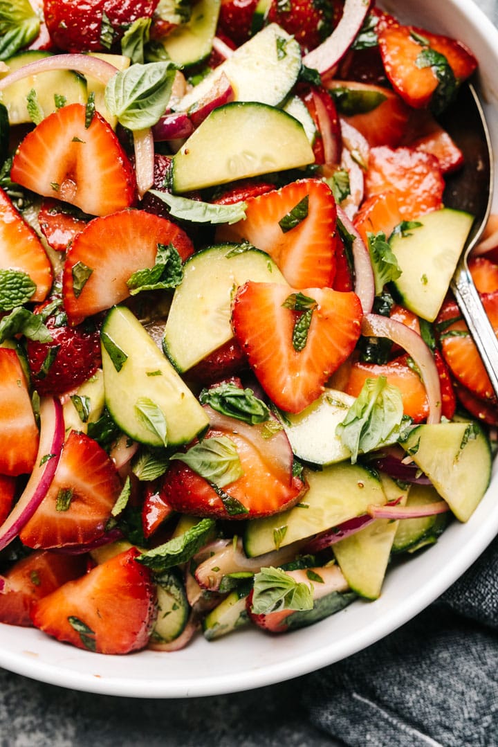 Detail from overhead, strawberry cucumber salad in a white mixing bowl with a vintage silver serving spoon.