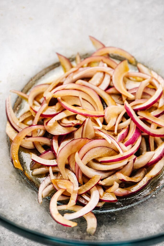 Thinly sliced red onions macerating in balsamic vinegar in a glass mixing bowl.