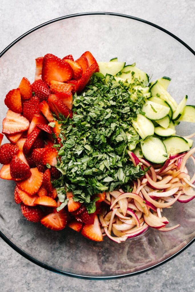 Sliced strawberries, cucumbers, red onions topped with minced fresh mint and basil in a glass mixing bowl.