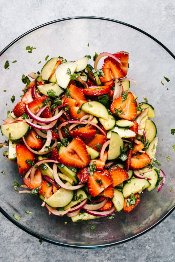 Tossed strawberry and cucumber salad with red onions and balsamic dressing in a glass mixing bowl.