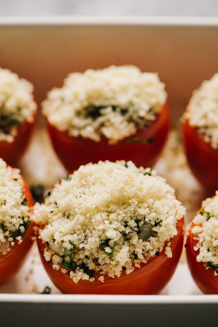Side view, tomatoes stuffed with quinoa filling and topped with cheese and breadcrumbs in a baking dish.