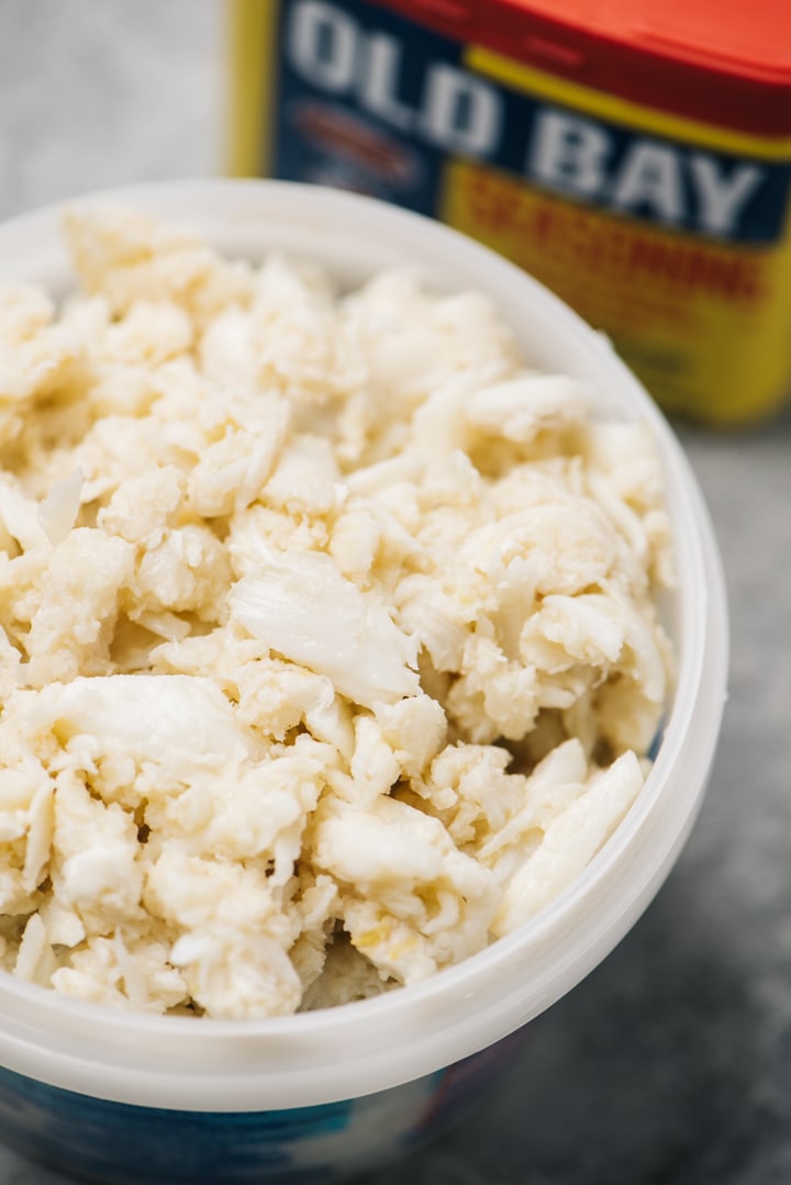 Side view, lump crab meat in a container with a box of old bay seasoning in the background.