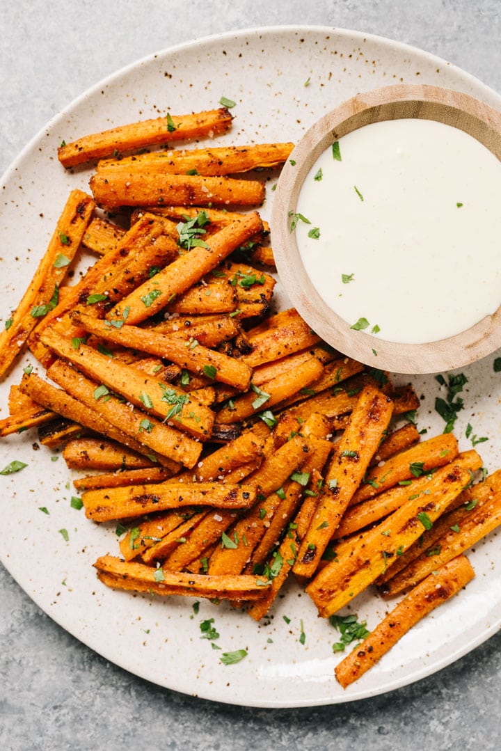 Carrot fries garnished with parsley and salt on a cream speckled plate with a side of garlic aioli dipping sauce.