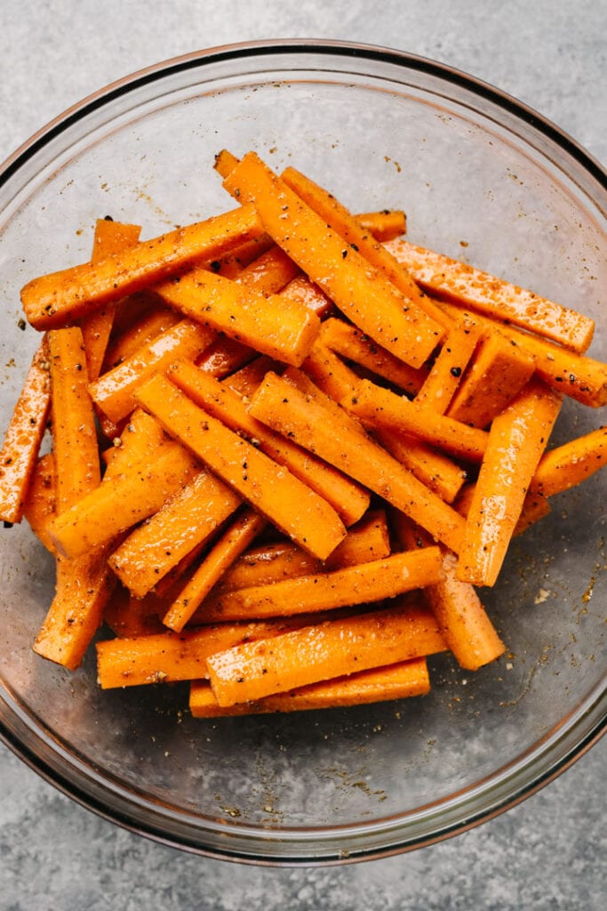 Carrot sticks tossed with olive oil and seasonings in a glass mixing bowl.