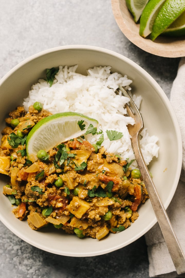 Keema curry over steamed white rice in a tan bowl with a fork and lime wedge.