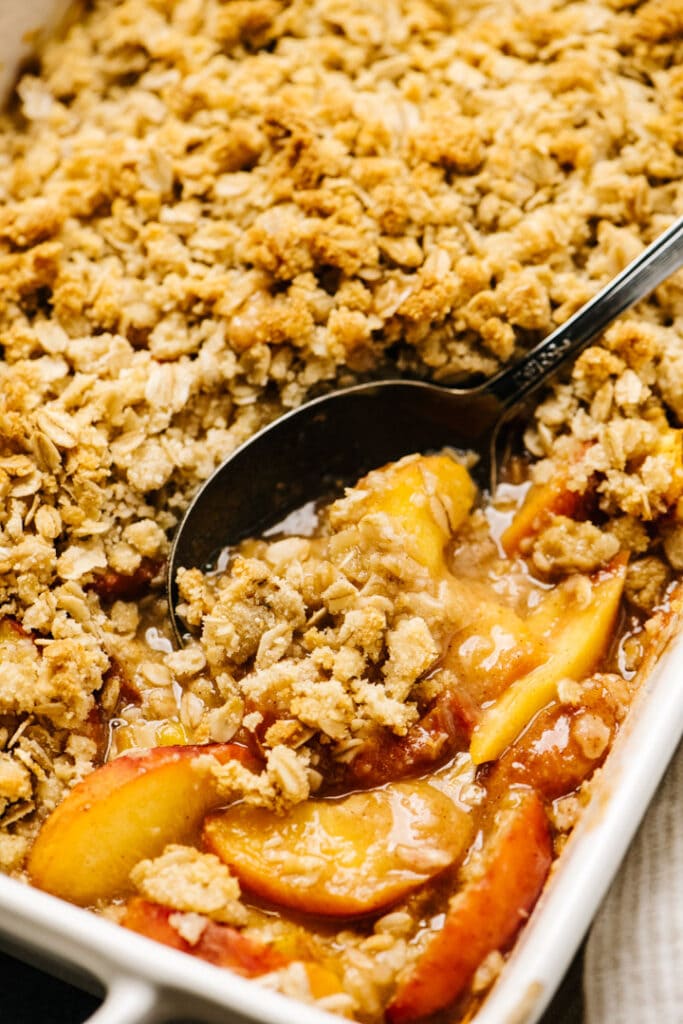 Side view, a serving spoon tucked into a casserole dish of warm peach crisp.