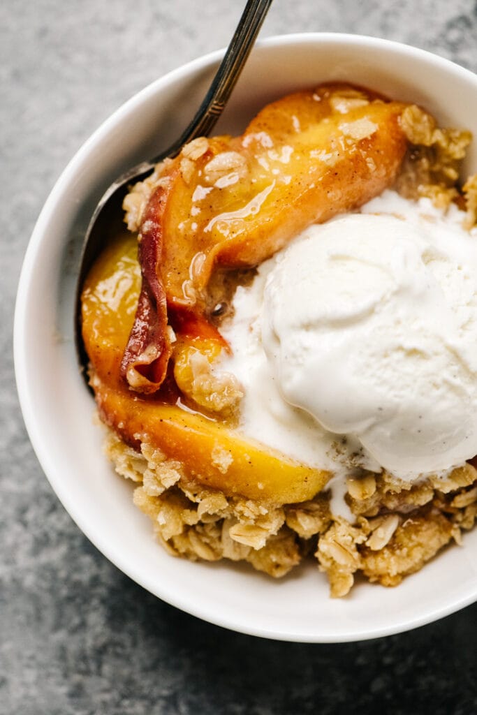 Healthy peach crisp in a white bowl topped with vanilla ice cream. 