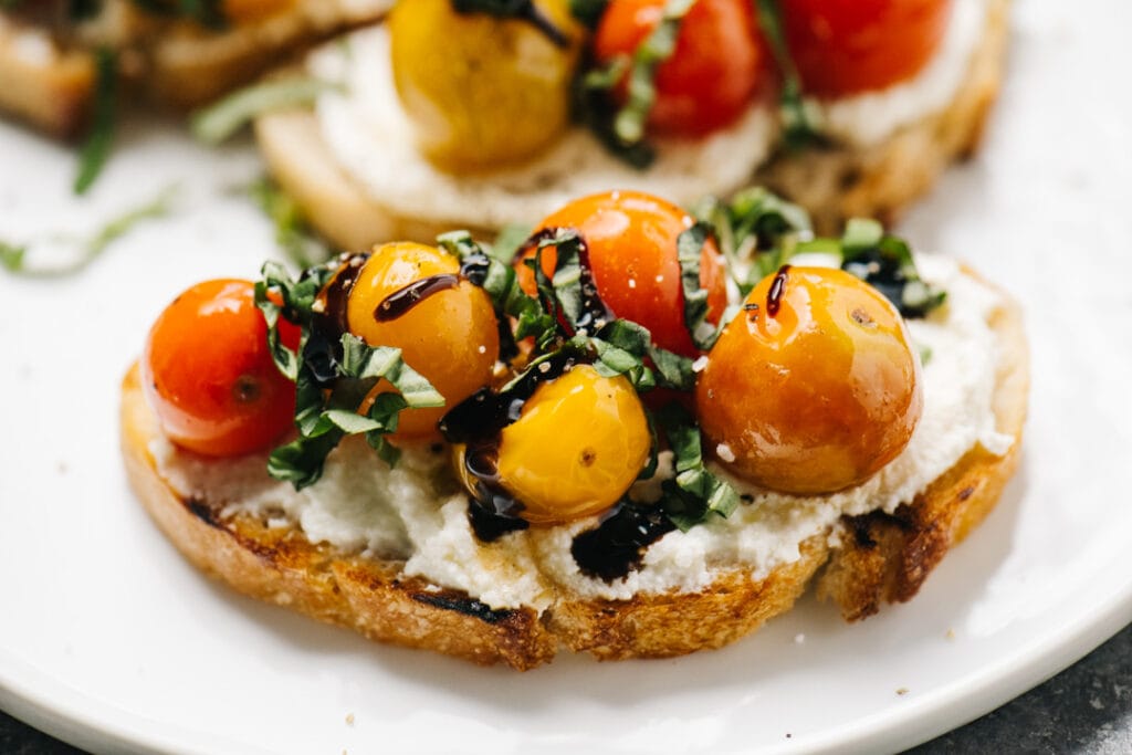 Side view, three pieces of ricotta toast on a white plate topped with tomatoes, basil, and balsamic glaze.