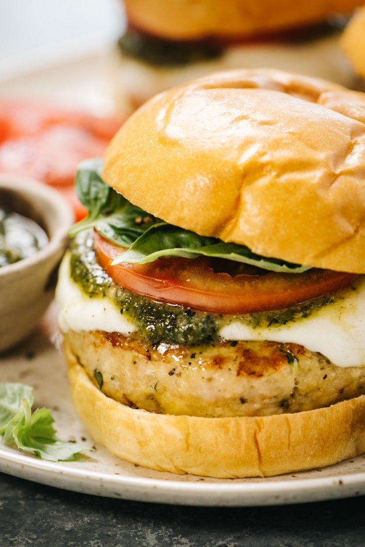 Side view, a caprese chicken burgers on a brown speckled plate made with chicken burgers, fresh mozzarella, pesto, tomato slices, and fresh basil leaves on a bun.