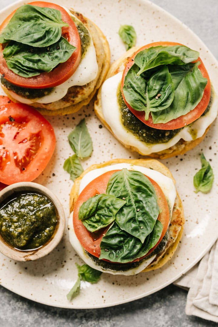Open faced caprese chicken burgers on a tan speckled plate with a small bowl of pesto, fresh basil, and tomato slices; cream linen napkin is tucked under the plate.
