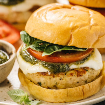 Side view, a caprese chicken burger on a plate made with a ground chicken burger topped with fresh mozzarella cheese, pesto sauce, sliced tomatoes, and fresh basil on a bun.