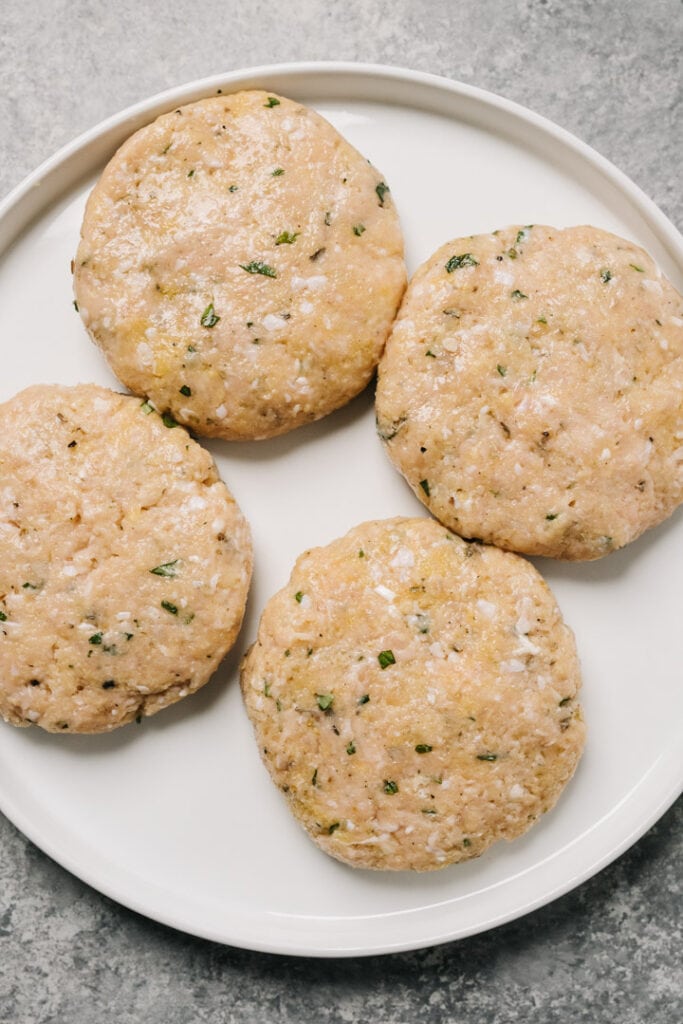 Four raw ground chicken burger patties on a white plate.