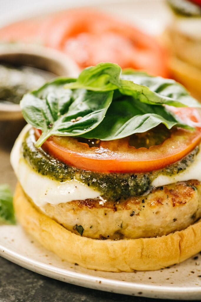 Side view, a ground chicken burger on a bun topped with fresh mozzarella cheese, pesto sauce, tomato slices, and fresh basil.