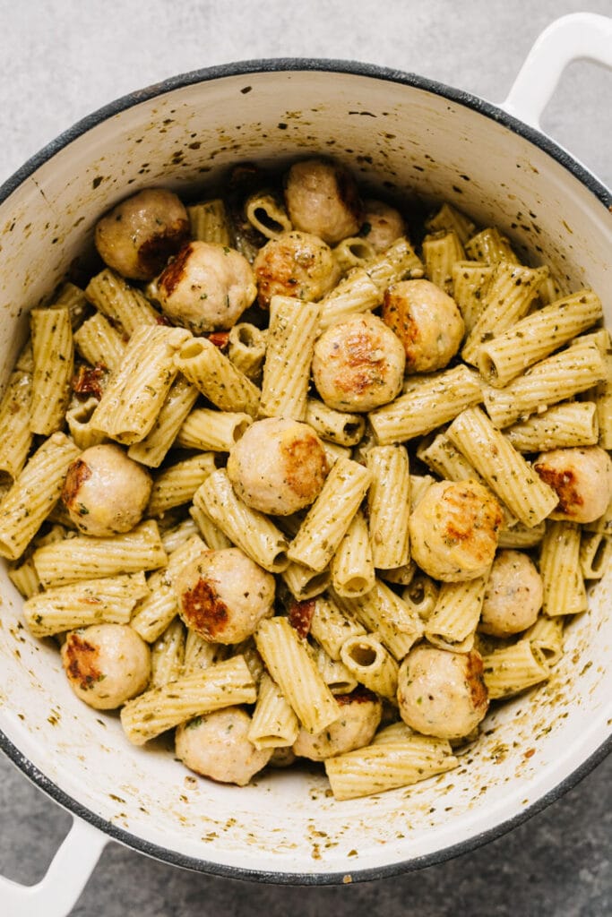 Chicken pesto pasta with meatballs and sun-dried tomatoes in a white pot.