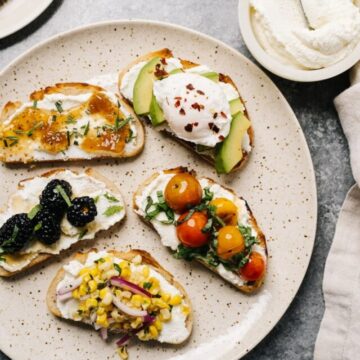 Five variations of ricotta toast on a cream speckled plate with a bowl of ricotta cheese on the side.