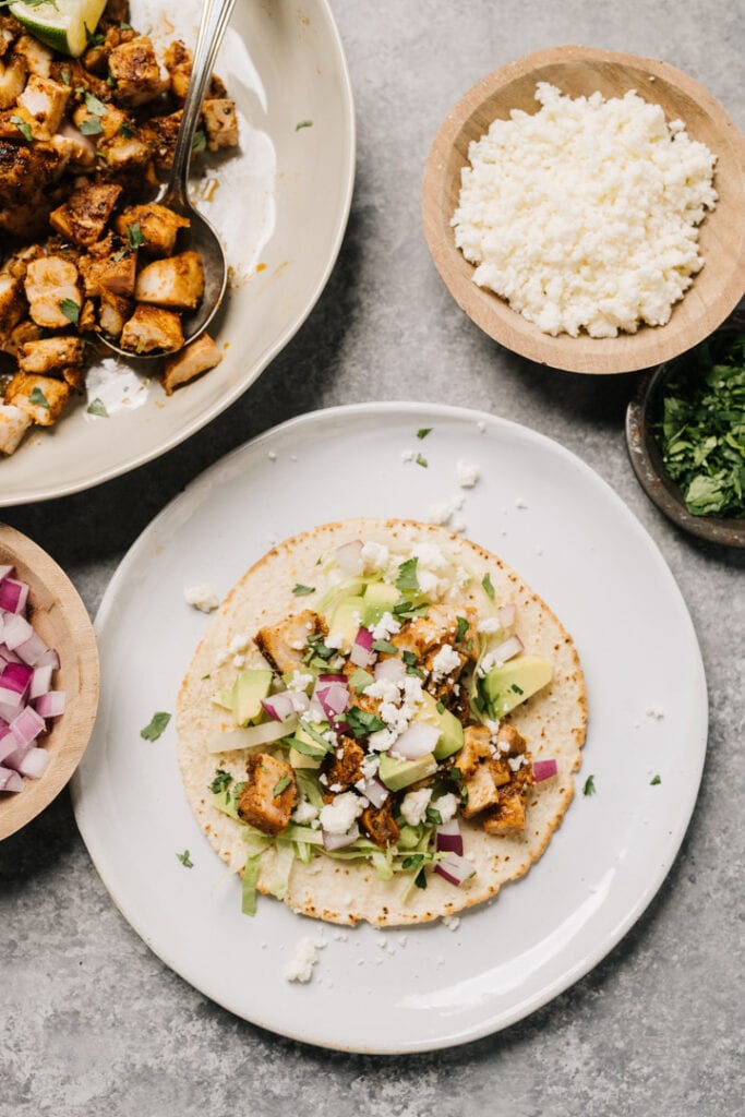 A grilled chicken taco in a corn tortilla served street-style, with red onion, cilantro, avocado, and queso fresco cheese.