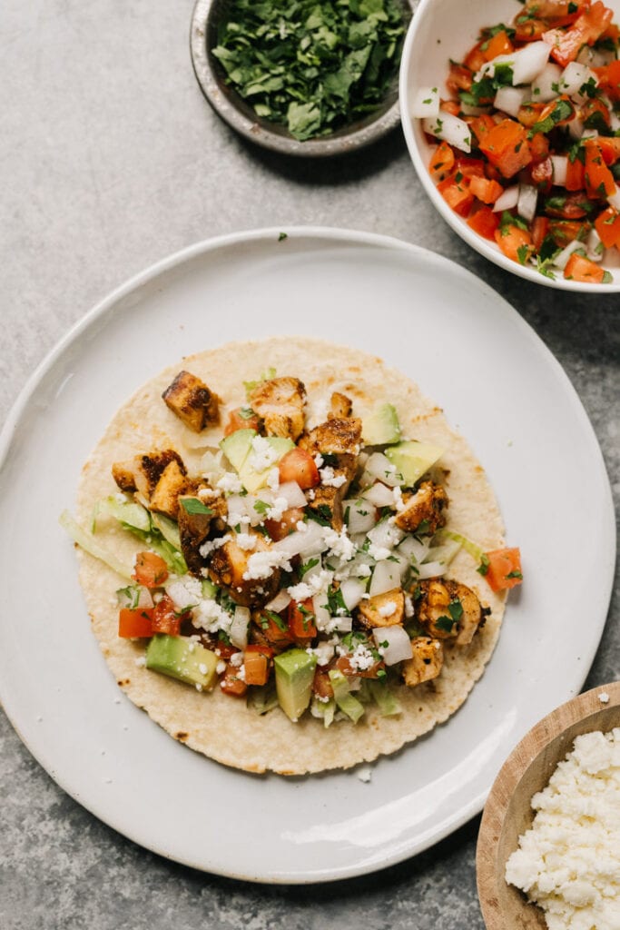 A grilled chicken taco in a corn tortilla served American-style with shredded lettuce, pico de gallo, avocado, and cheese.