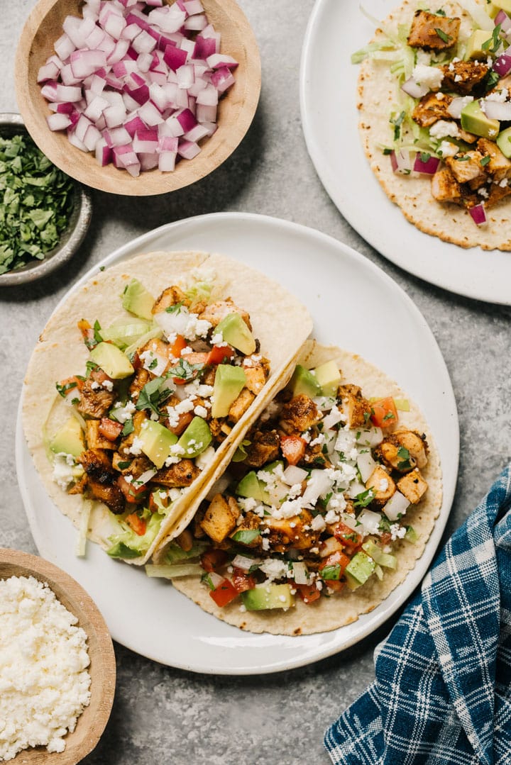 Two grilled chicken tacos on a plate, surrounded by small bowls of toppings and a blue linen napkin.