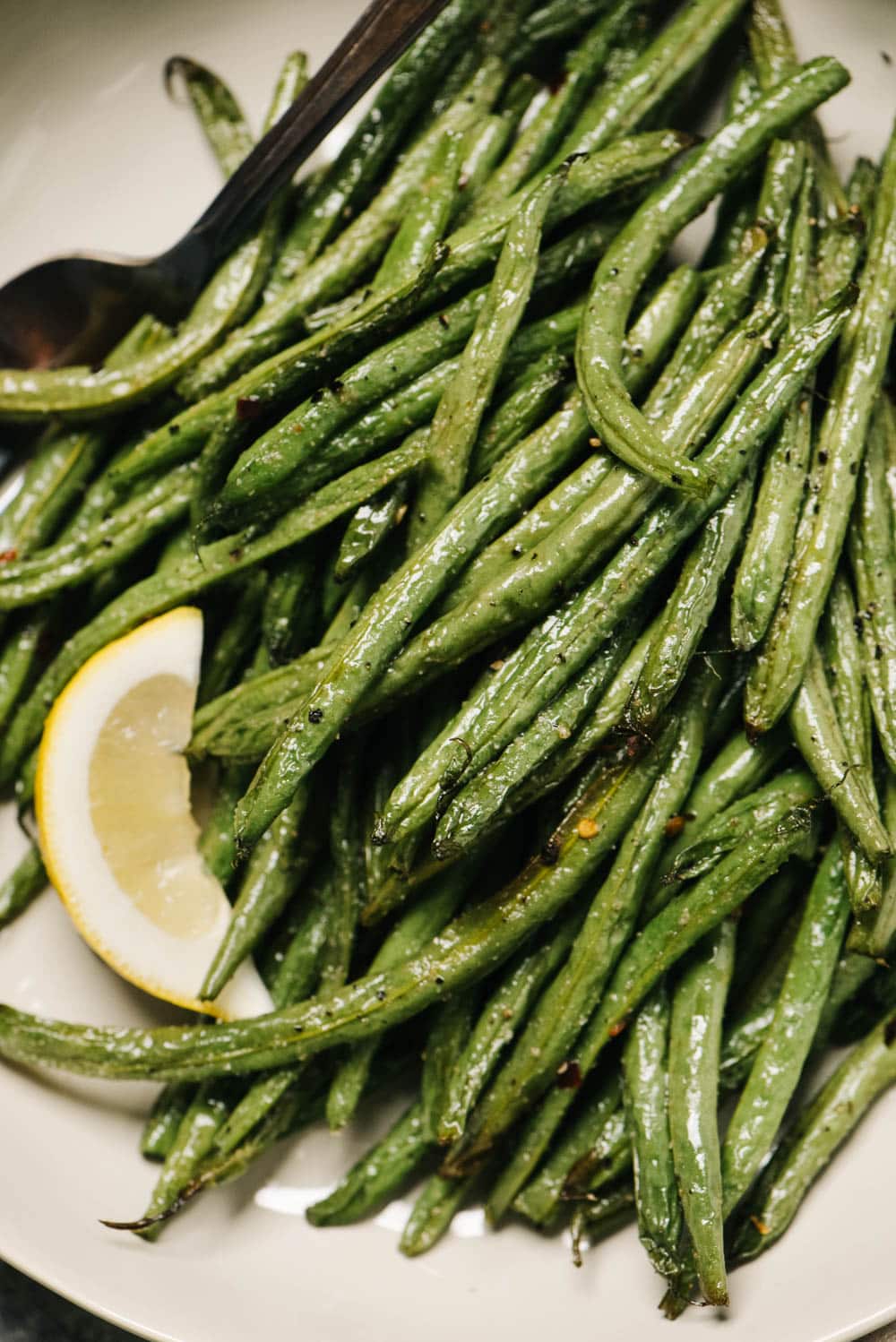Air fryer green beans in a tan serving dish with lemon wedges and a vintage serving fork.