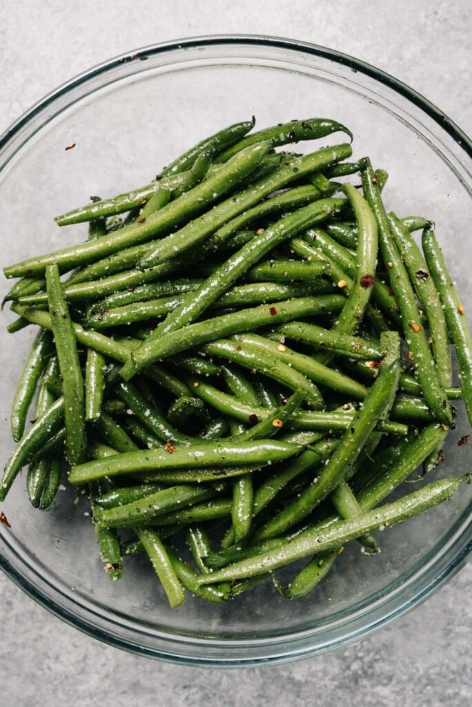 Fresh green beans in a glass mixing bowl, tossed with oil and seasonings.
