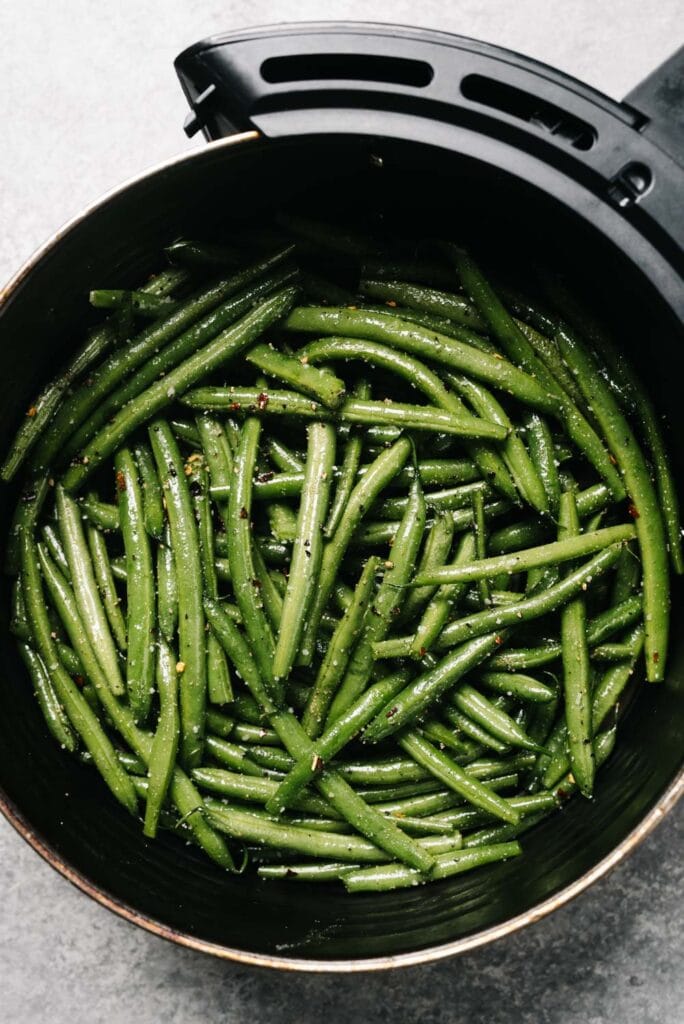 Green beans tossed with oil and seasoning in the basket of an air fryer.
