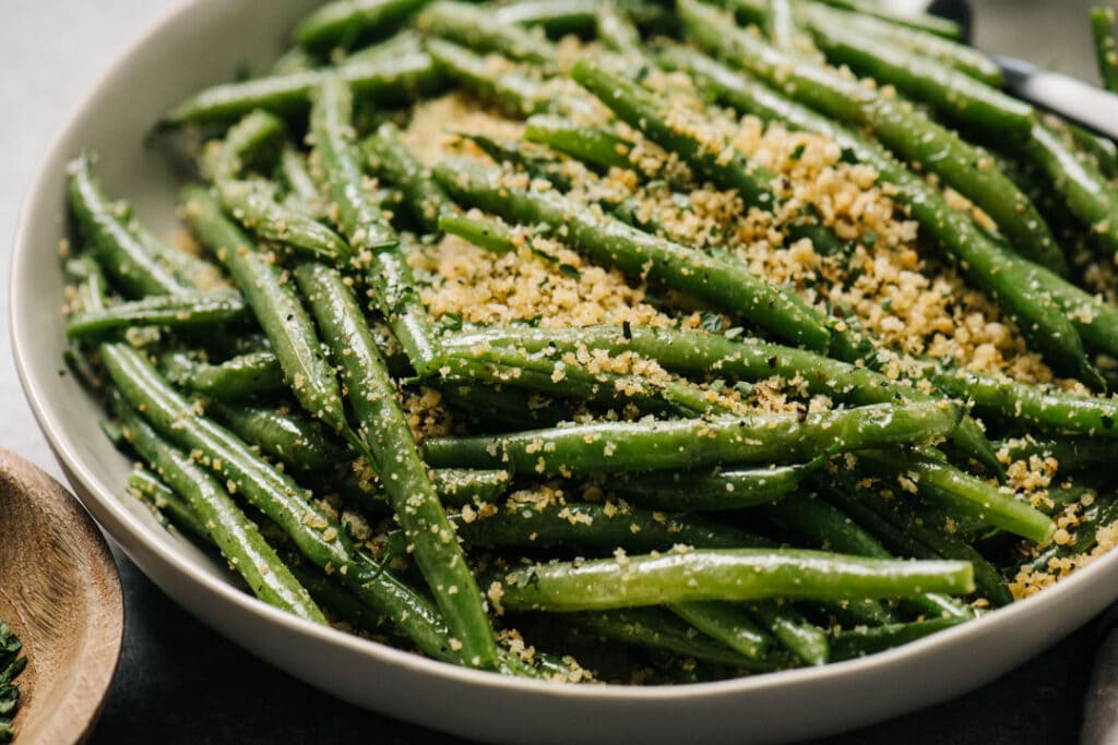 Side view, steamed green beans tossed with italian style breadcrumbs in a low tan serving bowl.