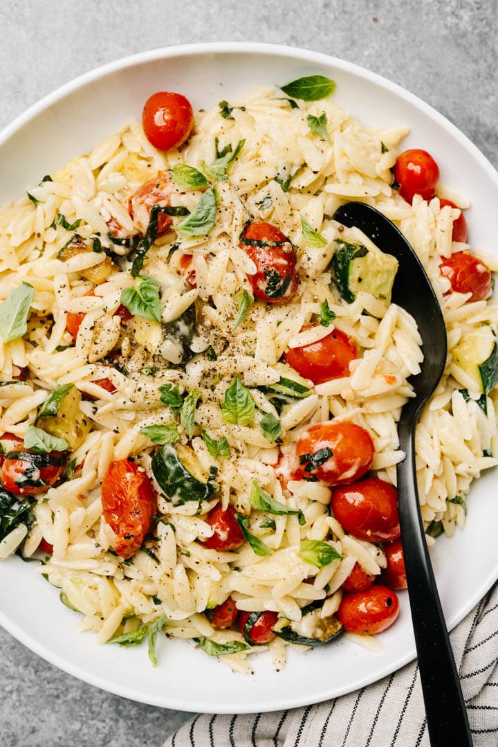 Orzo pasta recipe with tomatoes and zucchini in a large white serving bowl with a black spoon and striped linen napkin.