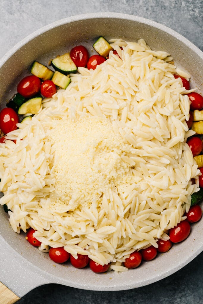 Sautéed vegetables in a skillet, topped with cooked orzo and grated parmesan cheese.