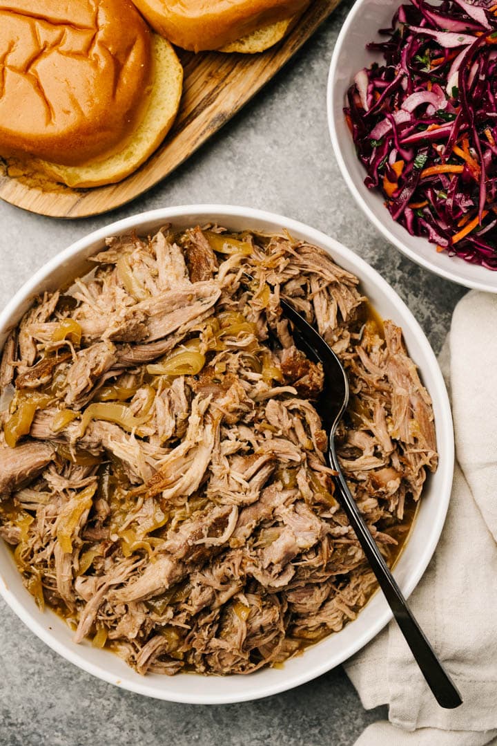 Cider pulled pork in a large white bowl surrounded with a bowl of coleslaw, brioche buns on a wood platter, and a tan linen napkin.