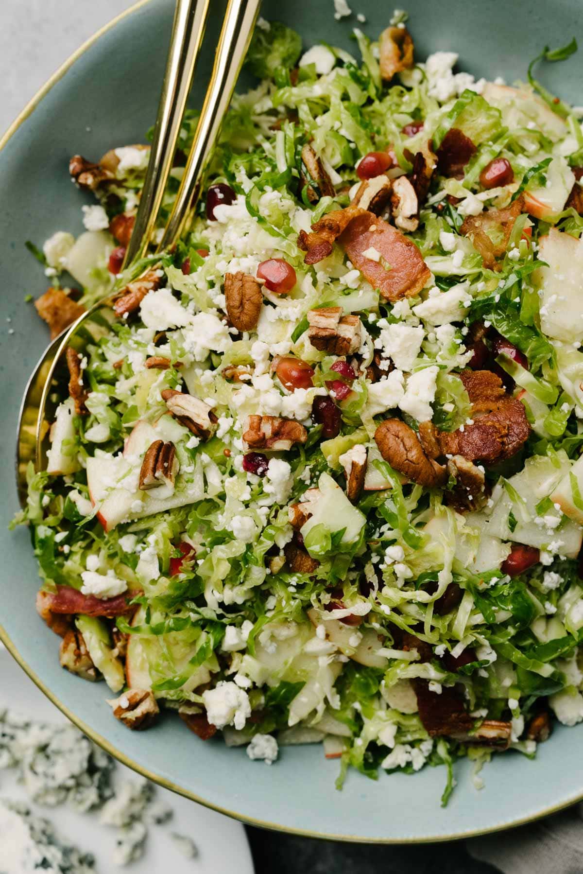 Gold serving utensils tucked into a Brussels sprout salad in a large blue, garnished with pecans, pomegranate seeds, and crumbled blue cheese.