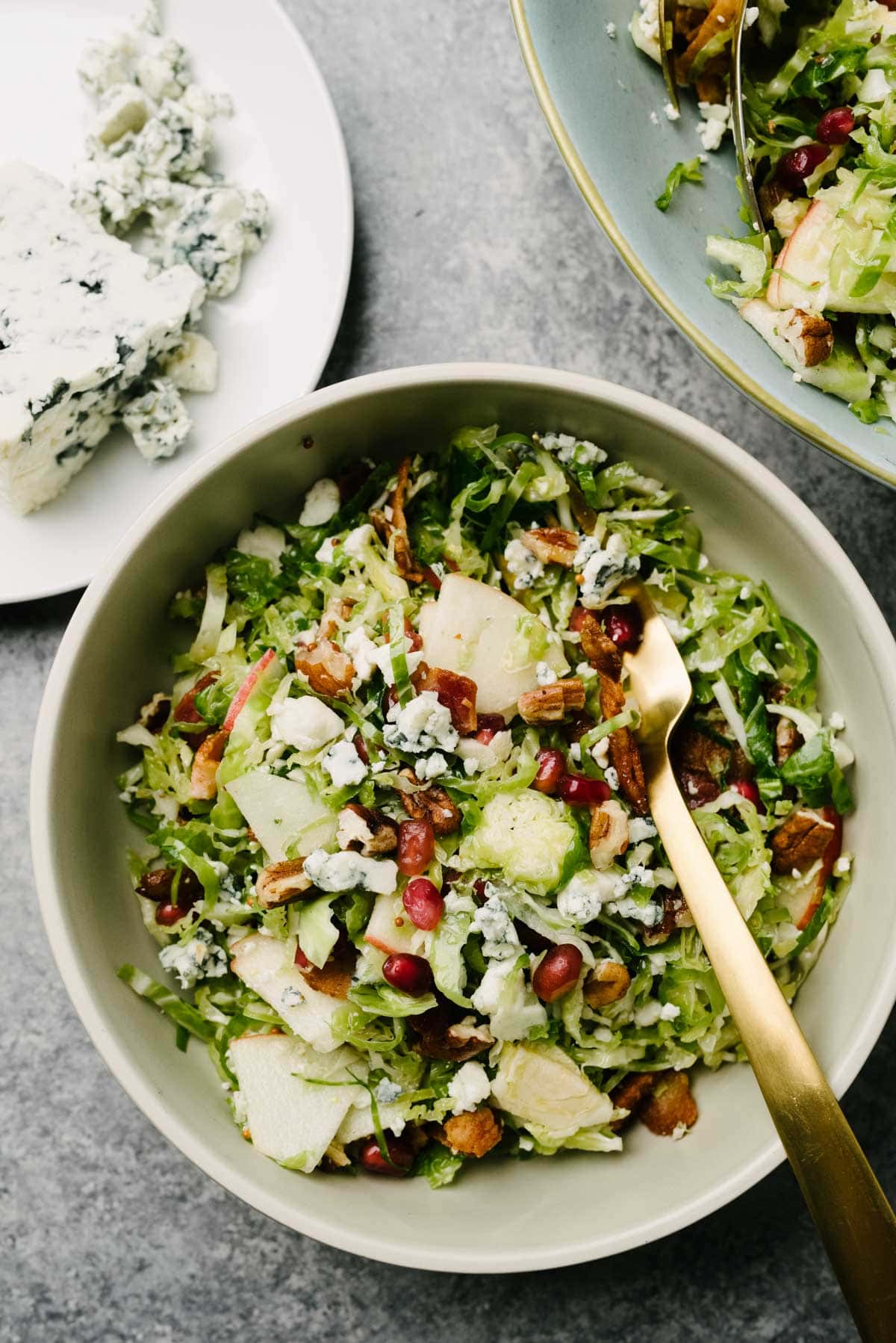 Brussels sprouts salad in a low tan salad bowl with a gold fork; a plate of blue cheese crumbles to the side.