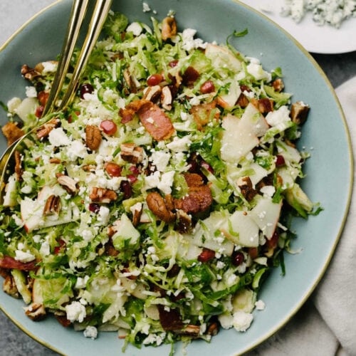 Brussels sprouts salad with bacon, apples, pomegranate seeds, pecans, and blue cheese in a blue salad bowl with gold serving utensils.