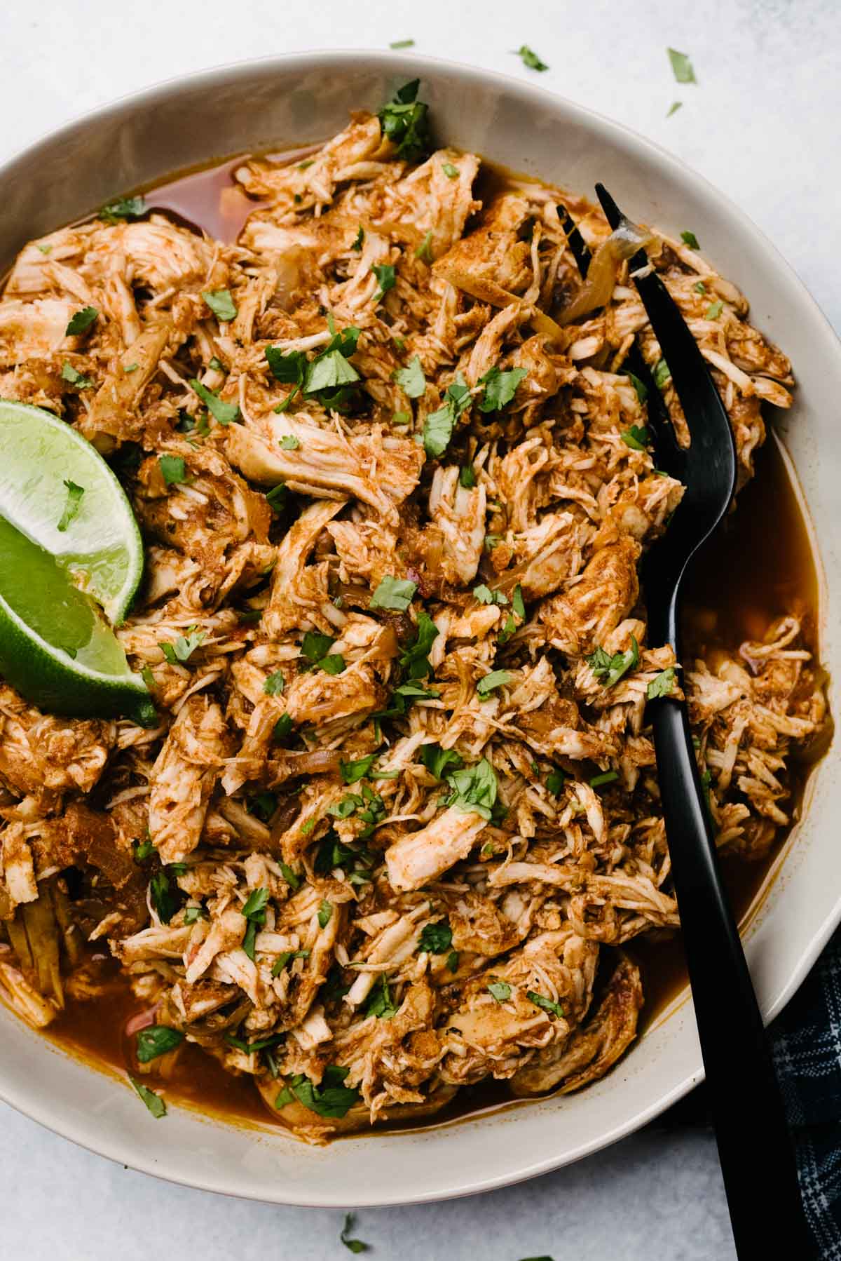 A black fork tucked in a bowl of crockpot shredded chicken taco meat, garnished with lime wedges and fresh cilantro.