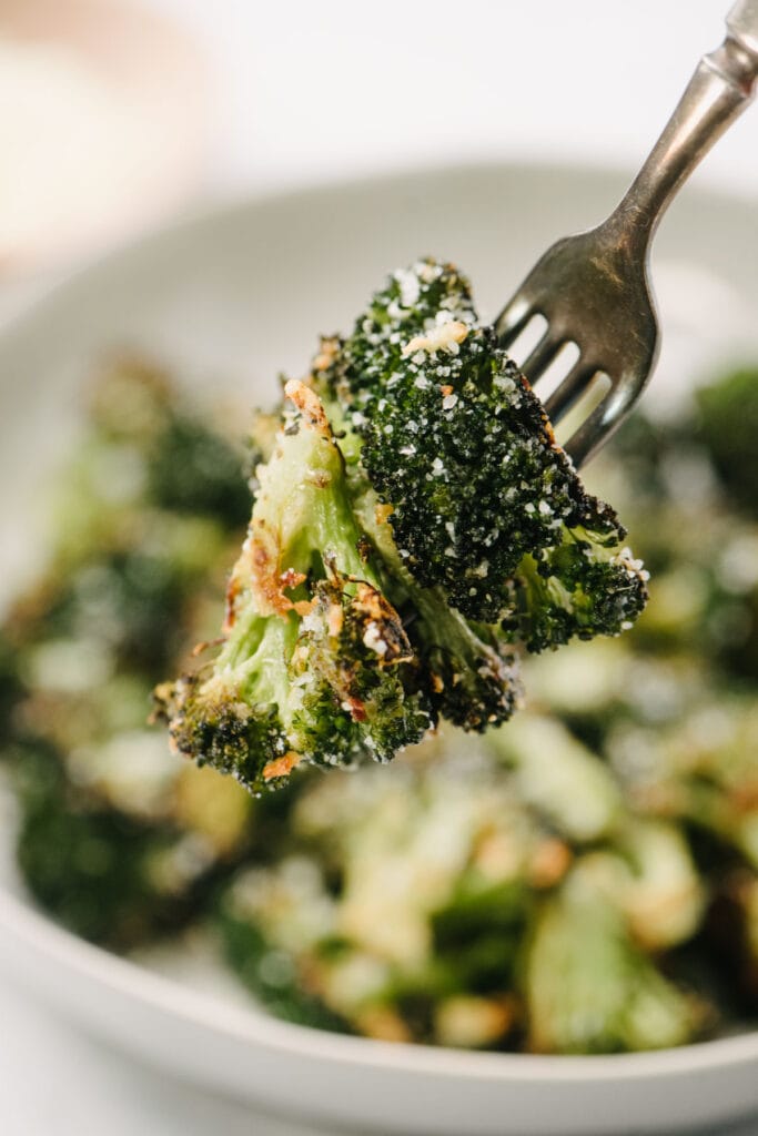 Two florets of roasted broccoli with parmesan on a fork hovering over a serving bowl.