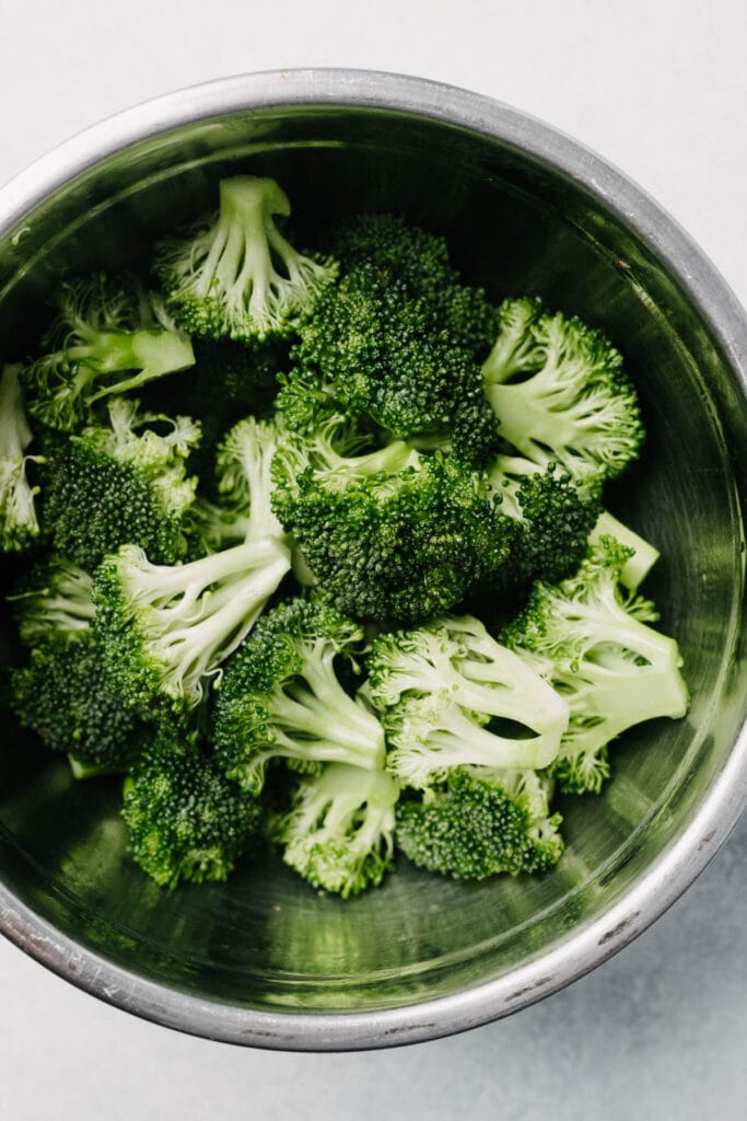 Broccoli florets tossed with olive oil in a metal mixing bowl.