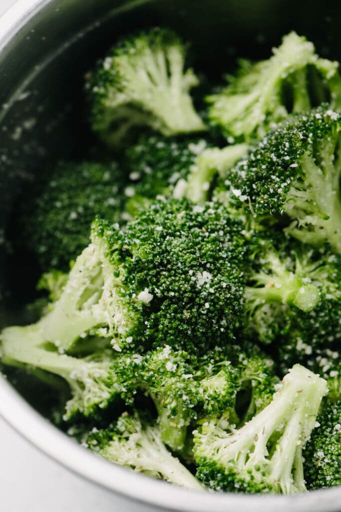 Broccoli florets tossed with olive oil, Italian seasoning, garlic powder, and parmesan cheese in a metal mixing bowl.