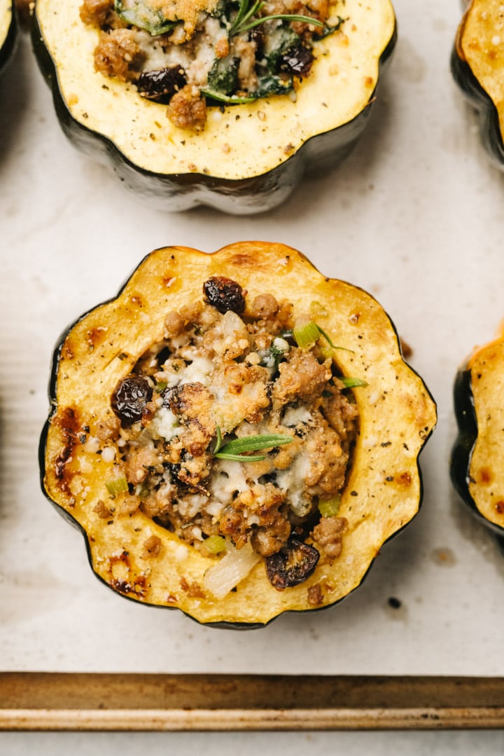 Several roasted acorn squash halves stuffed with sausage, vegetables, and fresh herbs and topped with grated parmesan cheese, arranged on parchment lined baking sheet.