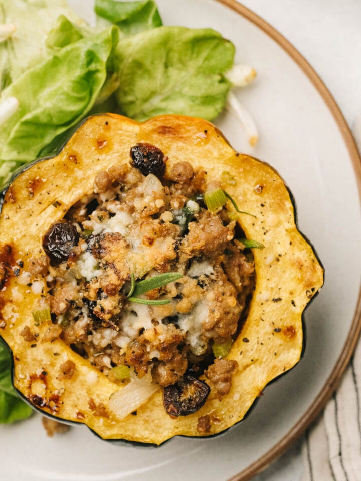 An acorn squash half stuffed with cooked sausage mixture, resting on a leaf of lettuce on a natural white colored plate.