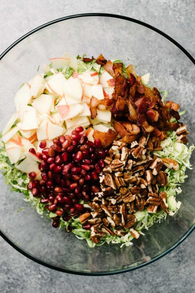 Shaved brussels sprouts apples, pomegranate seeds, bacon, and pecan in a large glass mixing bowl.