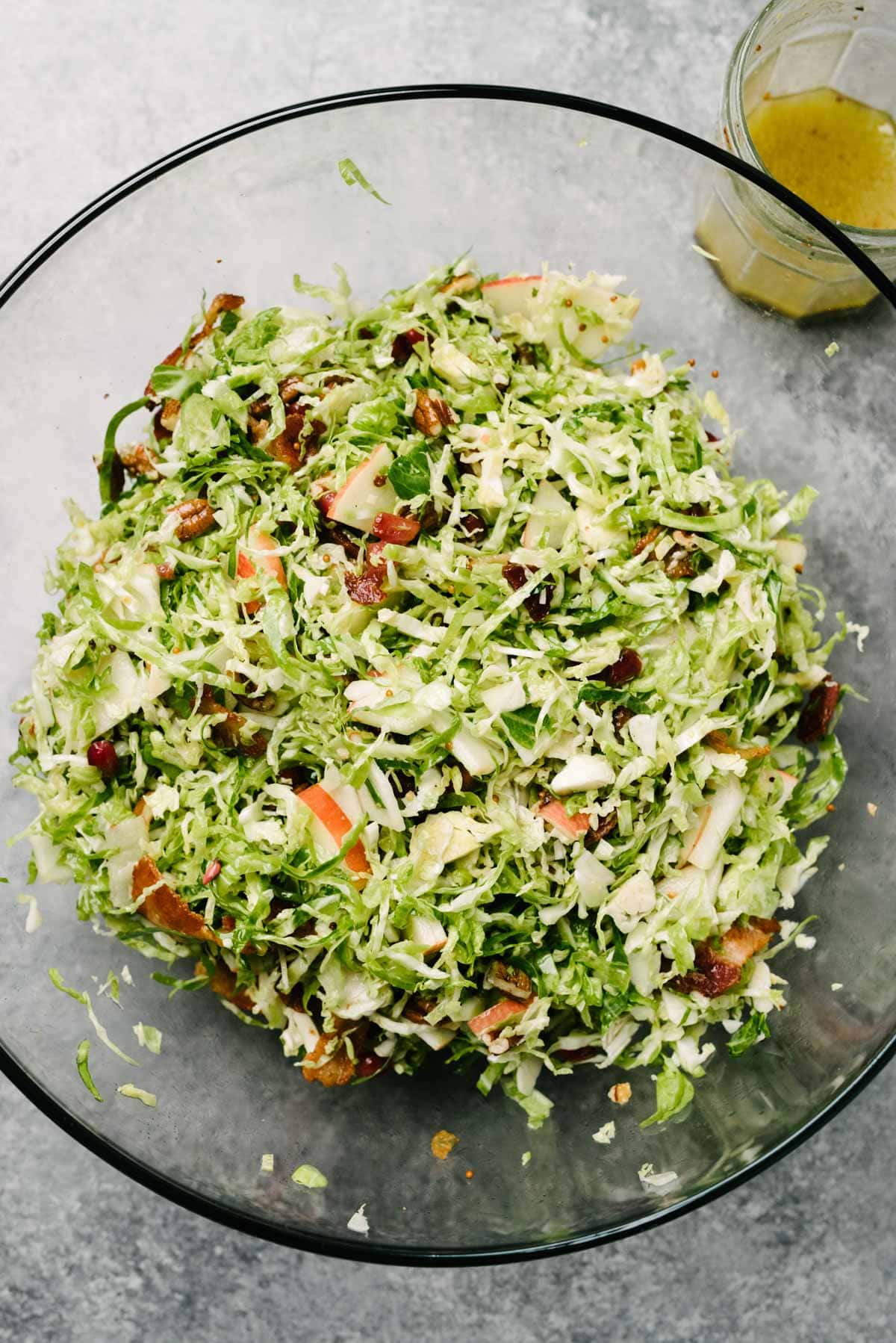 Tossed Brussels sprouts salad with bacon and apples in a large glass mixing bowl, with a small jar of vinaigrette to the side.