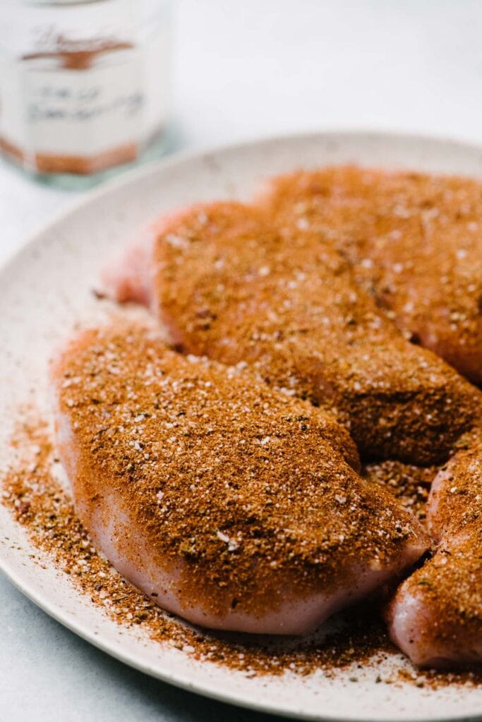 Raw chicken breasts on a plate topped with seasonings.