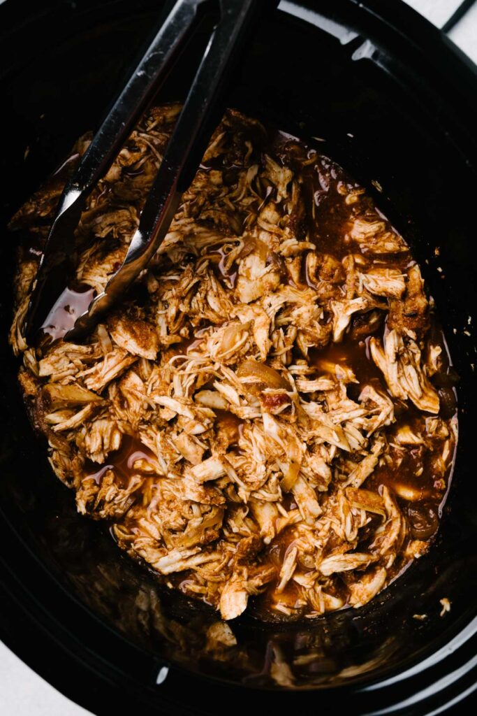 Tongs tucked into shredded chicken taco meat in a slow cooker.