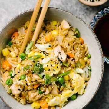 A bowl of turkey fried rice with a pair of chopsticks on a cement background, with small bowls of soy sauce and sesame seeds to the side.