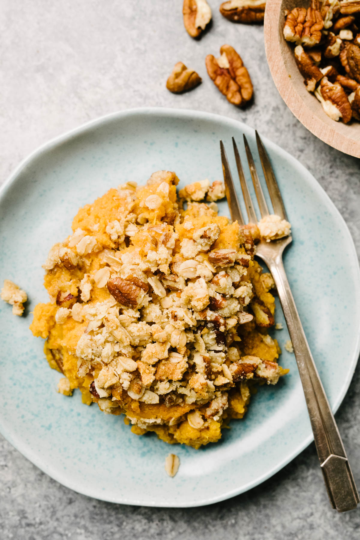 A light blue plate with a helping of sweet potato casserole topped with pecan streusel with a fork on the plate, and whole pecans on a side dish. 