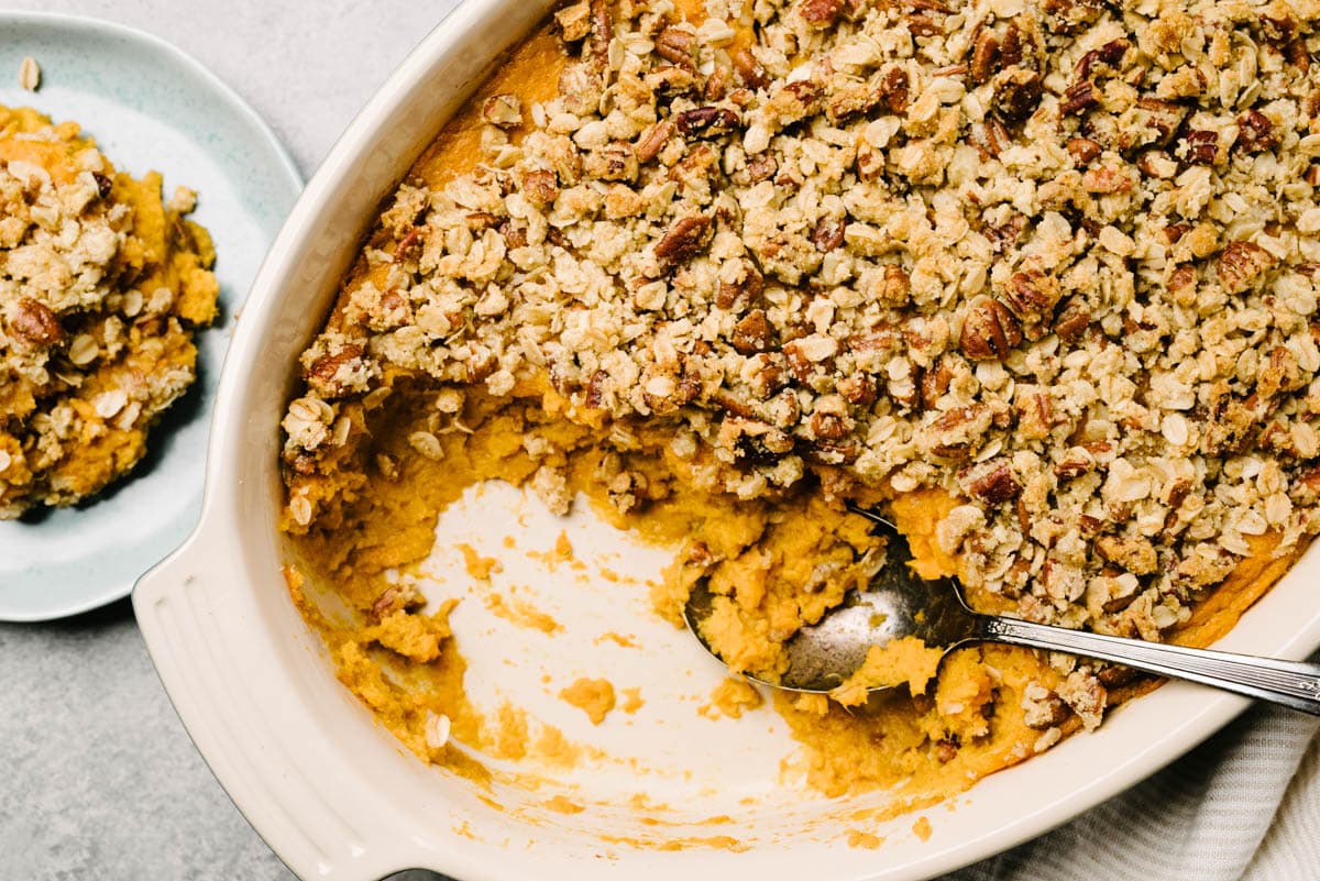 A baking dish with sweet potato casserole, with some served showing the layer of mashed sweet potatoes topped with a pecan streusel topping. 