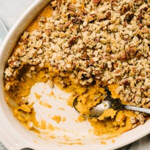 A baking dish of sweet potato casserole with pecan topping, with some scooped out to show the layers of mashed sweet potato topped with pecans.