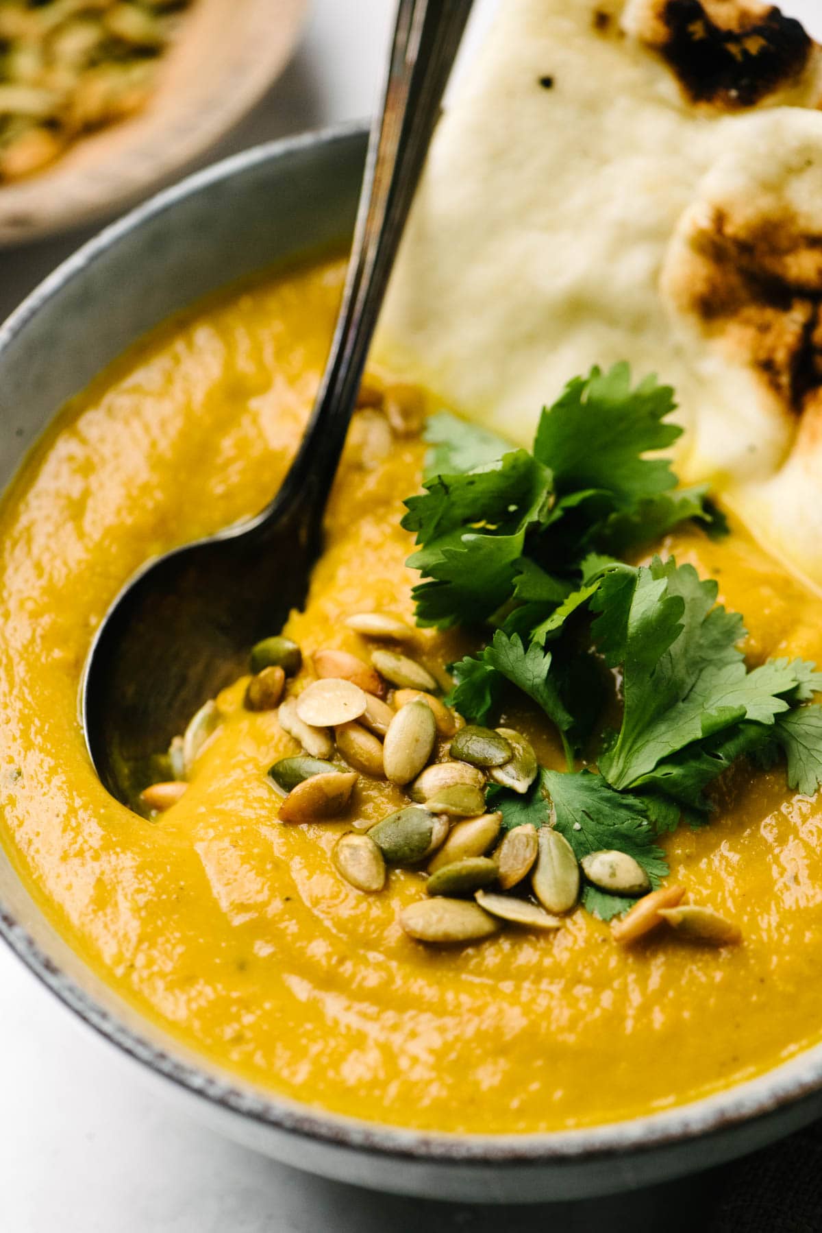 Side view, a spoon tucked into a bowl of pumpkin soup garnished with peptias and fresh cilantro.