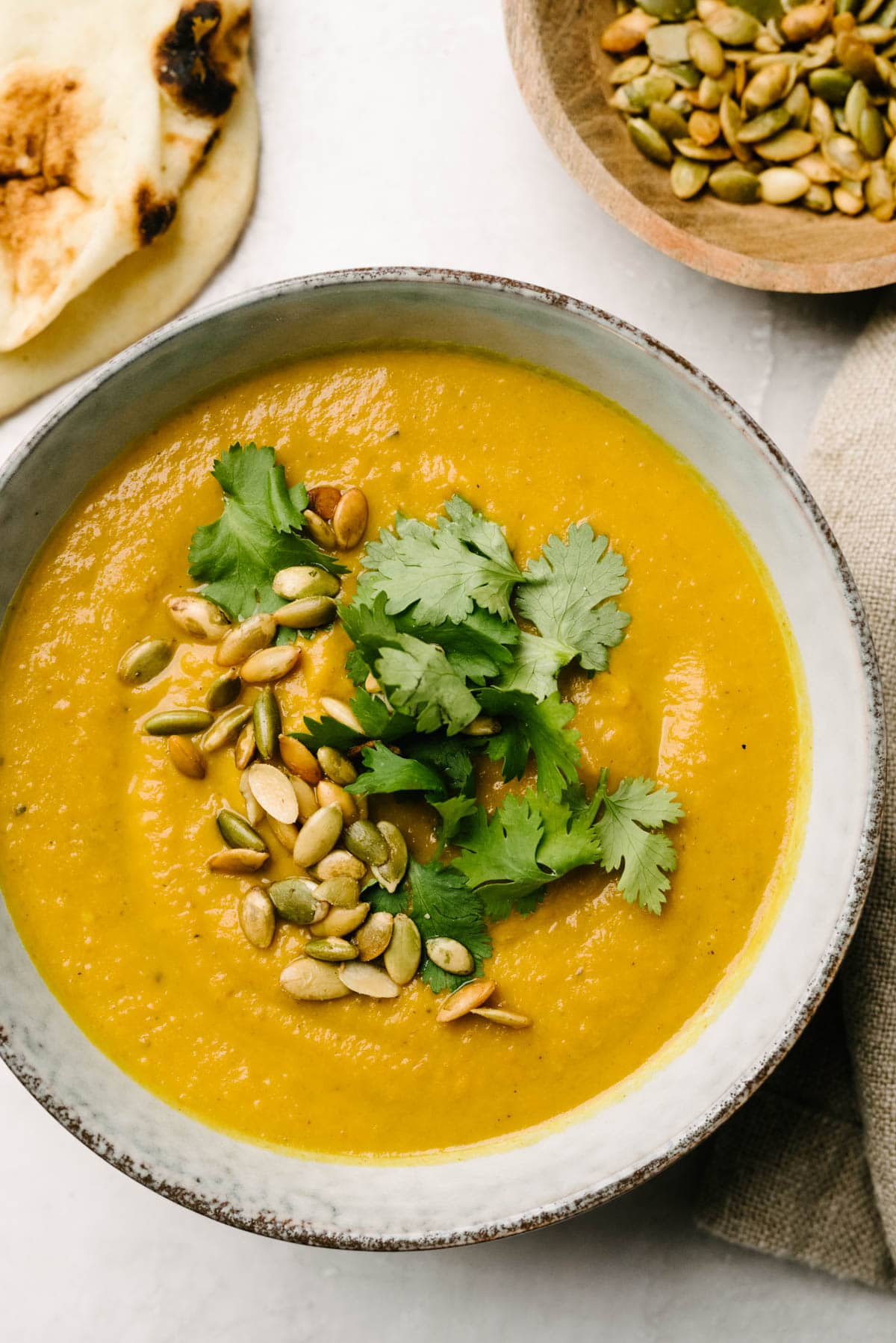 A bowl of pumpkin curry soup topped with pepitas and cilantro, with naan bread and a bowl of pumpkin seeds to the side.